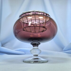 Puede incluir: Un cuenco de vidrio antiguo, color amatista, con un borde plateado y una base decorativa transparente. El cuenco tiene forma redonda y un inserto de rejilla met&aacute;lica. El vidrio es transparente, permitiendo el paso de la luz.