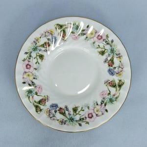 Peut inclure: Soucoupe en c&eacute;ramique blanche avec un motif floral. La soucoupe a un bord dor&eacute; et un motif en spirale. Le motif floral comprend des fleurs roses, jaunes, bleues et vertes.