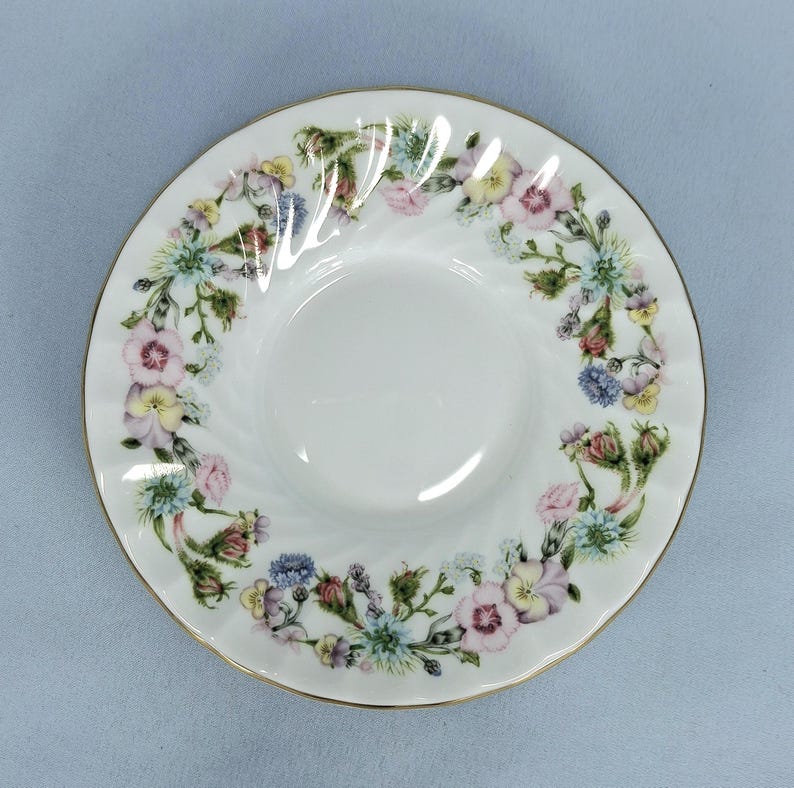 Peut inclure: Soucoupe en c&eacute;ramique blanche avec un motif floral. La soucoupe a un bord dor&eacute; et un motif en spirale. Le motif floral comprend des fleurs roses, jaunes, bleues et vertes.