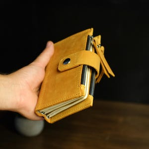 May include: A mustard yellow leather wallet featuring a snap button closure strap, a side zipper with a long leather pull tab, and multiple internal compartments visible through the side opening, showcasing a matte texture and stitched edges.