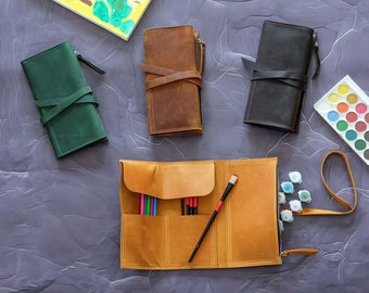 Può includere: Una vista dall'alto di materiali artistici su una superficie strutturata. In alto c'è un disegno di auto per bambini. Sono presenti diverse custodie in pelle verde, marrone e nera. Una custodia arrotolabile in pelle marrone chiaro contiene matite colorate e un pennello. È presente anche una tavolozza di acquerelli.