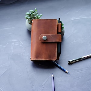 May include: A brown leather journal with a pen holder and a silver button clasp. The journal is closed and has a strap to secure it. A black pen is in the pen holder. A small plant, a blue pencil, and a patterned pen are also visible.