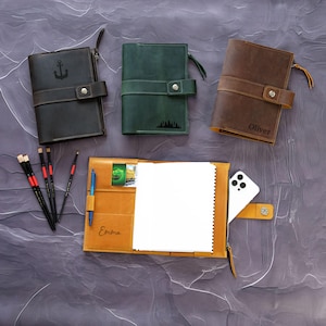 May include: Three leather-bound journals with personalized names and designs. The journals have a zipper closure and a flap with a button closure. The journals are on a gray textured surface.