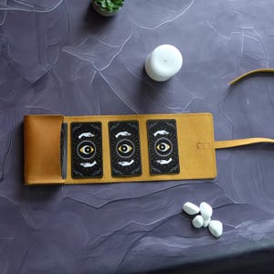 May include: A brown leather tarot card carrying case with three cards inside. The cards are black with a yellow eye and white hands. The case has a flap that closes with a snap.