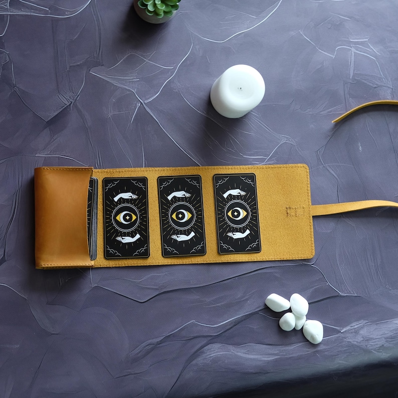 May include: A brown leather tarot card carrying case with three cards inside. The cards are black with a yellow eye and white hands. The case has a flap that closes with a snap.