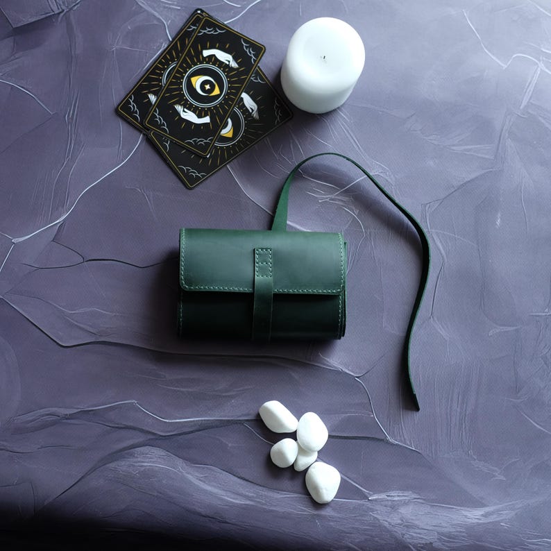May include: A dark green leather pouch with a strap, a white candle, and white stones on a purple textured surface. Two tarot cards with an eye and hands design are visible in the background.