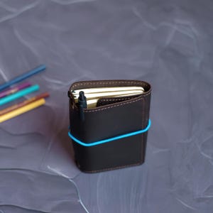 May include: A dark brown leather notebook cover with a blue elastic band. A black pen is inserted into the cover's pocket. The notebook is filled with cream-colored pages. Colored pencils are visible in the background.