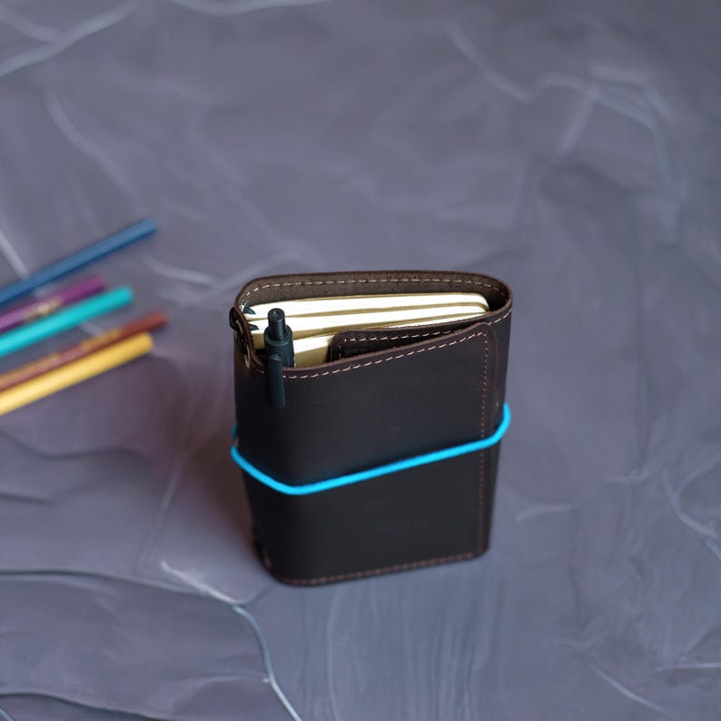 May include: A dark brown leather notebook cover with a blue elastic band. A black pen is inserted into the cover's pocket. The notebook is filled with cream-colored pages. Colored pencils are visible in the background.