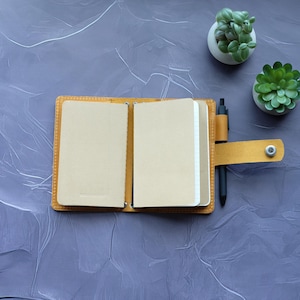 May include: Open, tan leather notebook with a pen holder and a black pen. The notebook has blank pages. Two small potted succulents are in the background on a textured, purple surface.