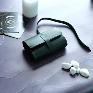 May include: A dark green leather wallet with a strap closure. The wallet is lying on a light gray surface with white stones.