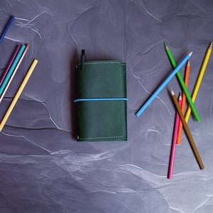 May include: A dark green leather journal with a blue elastic band and a black pen. Assorted colored pencils in yellow, blue, green, orange, pink, and brown are scattered around the journal on a gray surface. Stationery supplies.