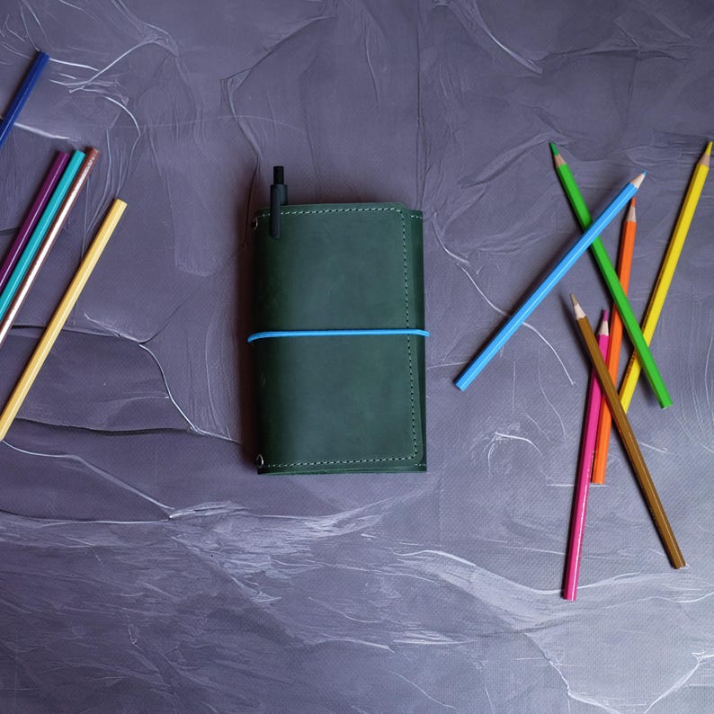 May include: A dark green leather journal with a blue elastic band and a black pen. Assorted colored pencils in yellow, blue, green, orange, pink, and brown are scattered around the journal on a gray surface. Stationery supplies.