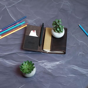 May include: Open brown leather journal with a black pen, a small succulent plant in a white pot, and a card in a pocket. The journal has the name "Oliver" embossed on the front. Colored pencils are scattered nearby.