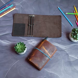 Könnte beinhalten: Ein braunes Lederjournal mit einem blauen Gummiband und einem Stift, neben einem offenen braunen Lederjournal. Buntstifte und kleine Topf-Sukkulenten sind ebenfalls zu sehen. Die Journale liegen auf einer grauen Oberfläche.