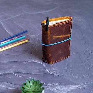 May include: A brown leather journal with a black pen inserted in the spine. The journal is secured with a blue elastic band. Colored pencils are visible in the background. A small green succulent plant is in the foreground.
