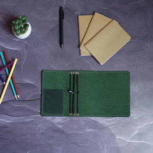 May include: A green leather journal cover with a small pocket and elastic closures, open to reveal the interior. A black pen, several small notebooks, and colored pencils are arranged on a textured purple surface, alongside a small potted succulent.