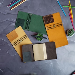May include: Several open leather journals in shades of green, tan, and brown. Each journal has elastic closures and internal pockets. Some journals are labelled with names such as "Mark", "Emma", and "Oliver". Pencils and a pen are also visible.