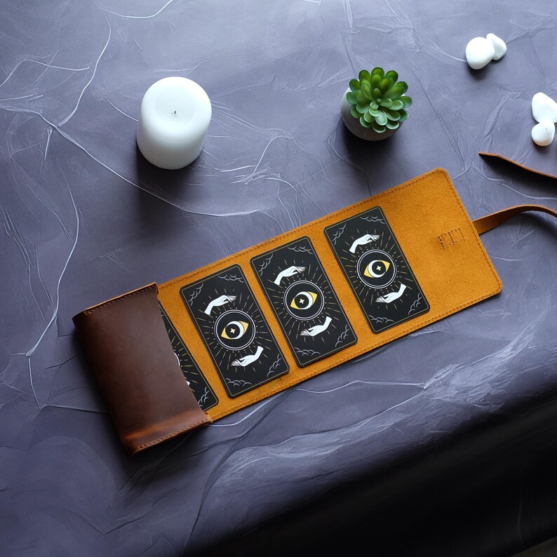 May include: A brown leather carrying case for tarot cards, unrolled to reveal five cards with a black background and a gold eye design. The cards are arranged in a row, with the eye facing forward on each card.
