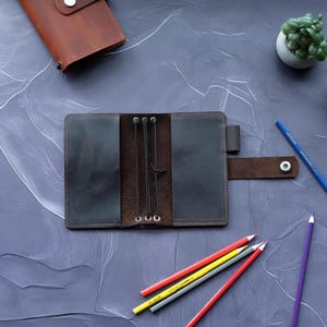 May include: Open, dark brown leather journal with elastic closures and a snap closure. A smaller, closed leather wallet is visible. Colored pencils in red, yellow, gray, blue, and purple are scattered around the journal. A small potted plant is also present.