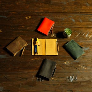 May include: Four leather notebook covers in red, brown, dark green, and dark gray, with one open yellow cover displaying a pen holder and two pens. Each cover features a side zipper and a rectangular shape for holding small journals.