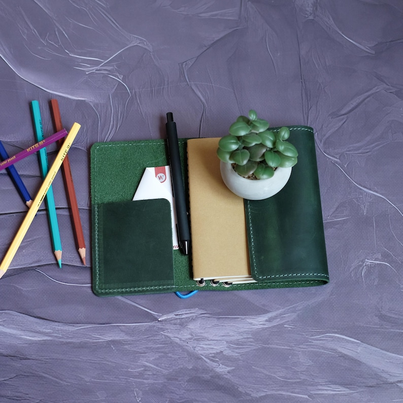 May include: A dark green leather journal with a pen, a small potted plant, and colored pencils. The journal is open, revealing a notepad and a card. The pencils are arranged on the left side of the journal.