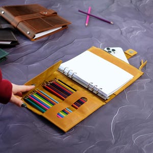 Pode incluir: Diário de couro amarelo mostarda aberto, com um bloco de notas branco e um conjunto de lápis de cor. Um smartphone está guardado no interior. Outros diários de couro em castanho e verde são visíveis no fundo.