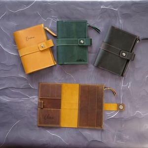 May include: A collection of leather journals in various colors: mustard yellow, forest green, and dark brown. Each journal has a strap closure and a zipper. One journal is open, revealing interior pockets and the name "Oliver". Another journal is labeled "Emma".