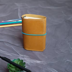 May include: A tan leather journal with a blue elastic band closure. The journal is rectangular with rounded edges and a stitched spine. A pen and a small succulent are in the foreground.
