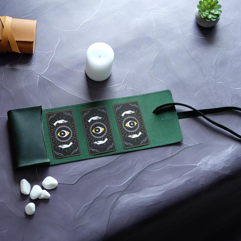 May include: A green leather tarot card carrying case with a strap. The case is open, revealing three cards with a black background and a gold eye design.