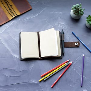 May include: An open, dark brown leather journal with blank pages, a pen, and a button closure. Colored pencils in red, yellow, gray, and purple are scattered around the journal. A second leather journal is visible in the background.