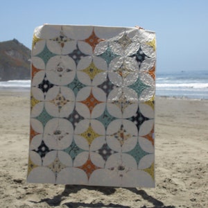 K&ouml;nnte beinhalten: Eine wei&szlig;e Steppdecke mit einem geometrischen Muster aus orangefarbenen, gelben, blauen und grauen Kreisen und Sternen. Die Steppdecke h&auml;ngt an einem Strand.