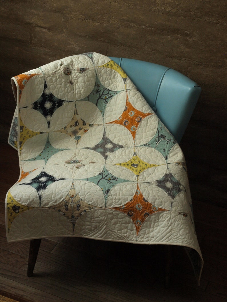 K&ouml;nnte beinhalten: Eine wei&szlig;e Steppdecke mit einem geometrischen Muster aus Kreisen und Sternen in Blaut&ouml;nen, Gr&uuml;n, Gelb und Orange. Die Steppdecke ist &uuml;ber einen blauen Sessel gelegt.