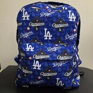 May include: Blue backpack featuring the Los Angeles Dodgers logo and the words "Champions" and "Dodgers" in white. The backpack has a repeating pattern of the team's logo and text, with baseballs scattered throughout.