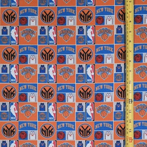 May include: A fabric print featuring a repeating pattern of New York Knicks logos, basketballs, and team jerseys in orange, blue, and white. A yellow measuring tape is visible on the right side, showing a length of approximately 16 inches.