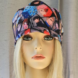 May include: A black beanie with a colorful anime print featuring a blue die with eyes, characters, and a comic book style background.
