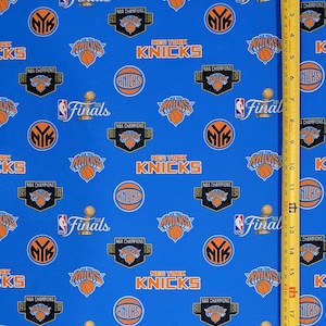 May include: A blue fabric featuring a repeating pattern of New York Knicks logos, NBA Finals 2026 graphics, and NBA Champions 2026 emblems in orange, white, and black. A yellow measuring tape is placed vertically, showing a length of 18 inches.