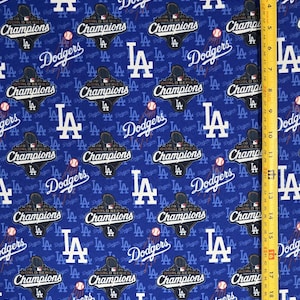 Puede incluir: Una tela azul con un patrón repetitivo de logotipos de los Los Angeles Dodgers, pelotas de béisbol y emblemas de 2020 World Series Champions. Una cinta métrica amarilla está colocada a la derecha, mostrando una longitud de 46 centímetros.