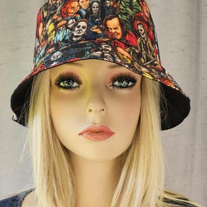 Puede incluir: Un sombrero de pescador reversible con un colorido collage de personajes de películas de terror. El sombrero tiene un ala negra y un interior negro. El diseño incluye imágenes de Michael Myers, Freddy Krueger y otras figuras de terror icónicas.