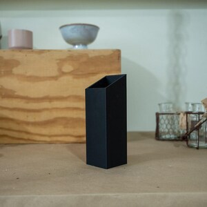 May include: A black, rectangular vase with an angled top sits on a light brown surface. The vase is the focal point, with other decorative items like bowls and small pots in the background.