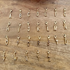 May include: Gold-colored alphabet charms, each with a lobster clasp, are arranged on a wooden surface. The letters A through Z are displayed, offering a personalized touch for jewelry or crafts. The charms are approximately 0.75 inches in height.
