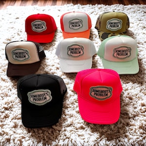 May include: A selection of trucker caps in diverse colours, such as red, orange, green, and black. Each cap has a patch with the text "SOMEBODY'S PROBLEM" in a stylized font. The caps are arranged on a textured surface.
