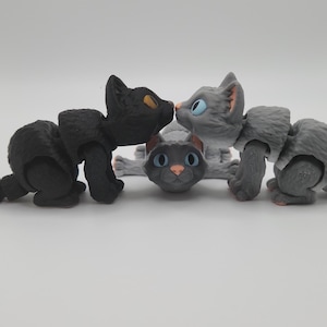 May include: Three articulated toy cats, one black, one gray, and one white with gray markings. The black cat is kissing the gray cat on the head.