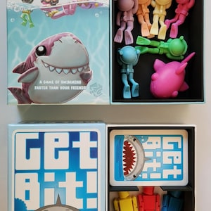 May include: A blue box with a shark on the front and the words "Get Bit!" in white. The box contains a game with colorful plastic figures and cards. The game is by Dave Chalker.
