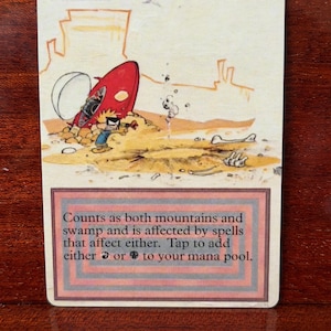 May include: A rectangular trading card titled Badlands featuring a cartoon illustration of a character in a blue shirt standing near a red spaceship in a desert landscape. The text reads: Counts as both mountains and swamp and is affected by spells that affect either. Tap to add either red or black mana to your mana pool.