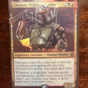 Op de afbeelding: Een Magic: The Gathering-kaart met "Chainer, Nightmare Adept". De kaart toont een personage in groene en bruine bepantsering met een helm. De kaarttekst bevat "Legendary Creature - Human Minion" en spelinstructies. De kaart heeft 3/2 kracht/weerstand.