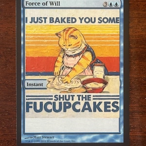 May include: A Magic: The Gathering card titled "Force of Will" featuring a cat baking cupcakes. The image includes the text "I JUST BAKED YOU SOME" and "SHUT THE FUCUPCAKES". The card has a blue border and the artist's signature.