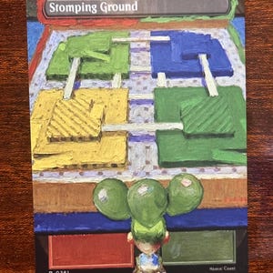 Puede incluir: Una tarjeta colorida con el texto "Stomping Ground" en la parte superior. La tarjeta tiene un diseño pintado con secciones amarillas, verdes y azules. Una pequeña figura con orbes verdes está en la parte inferior. La tarjeta también tiene el texto "R 0281 CLU'EN GIIGUIAN" y "Mimia Coast".