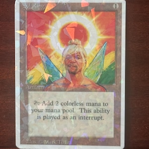 Peut inclure: Une carte à collectionner holographique Sol Ring illustrée d'une figure ailée sur un fond de soleil. Le texte indique Sol Ring, Artifact, et la capacité : Add 2 colorless mana to your mana pool. This ability is played as an interrupt. Illus. Mark Tedin.