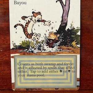 May include: A rectangular trading card titled Bayou featuring a cartoon illustration of a tiger and a child splashing in a muddy pool. The text reads: Counts as both swamp and forest and is affected by spells that affect either. Tap to add either black or green mana to your mana pool.
