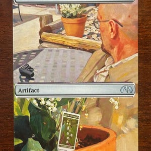 May include: A Magic: The Gathering card labelled "Lotus Petal" with a mana cost of zero. The card shows an artistic image of a man with glasses, a potted plant, and a small camera on a table. The card type is "Artifact".